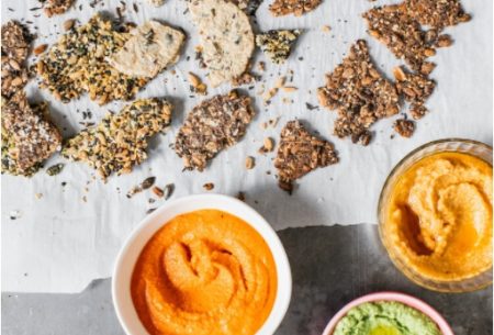 Two small pots of harissa red pepper dip, shown on bench top surrounded by mixed broken seed crackers