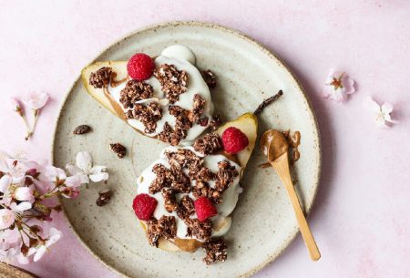 A plate with a pear halved with yogurt, nut butter, granola and nut butter on top with a spoon of nut butter on the side of the plate