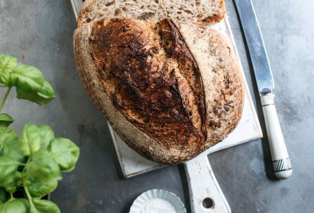 A loaf of sourdough that has been slices alongside a knife, salt, pepper, basil and a bottle of oil