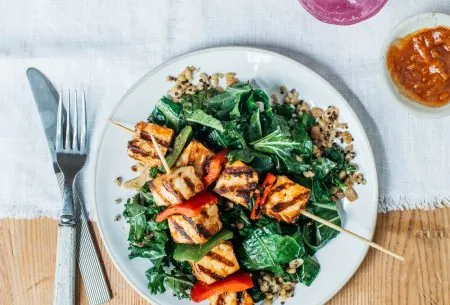 A plate with quinoa, kale and veggie skewers on a wooden table with a white linen cloth and knife and fork, pink drink with ice and red dip