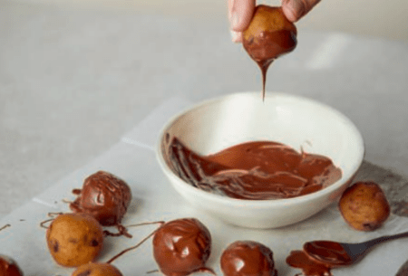 A small bowl of melted chocolate with a hand dipping in a cookie dough ball to cover in chocolate, other balls already covered in chocolate and not covered in chocolate are alongside in rows