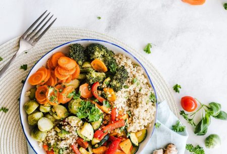 A colourful bowl of food including quinoa, roasted mixed peppers, courgette, carrot, brussell sprouts and broccoli on a place mat with a napkin and piece of ginger