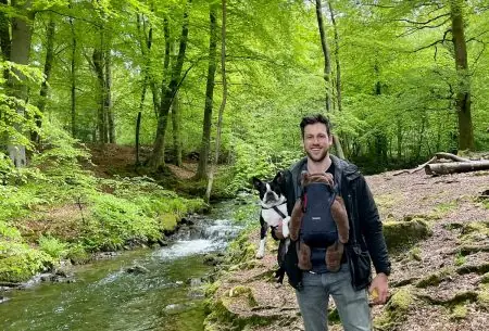 Man with baby strapped to chest, standing outdoors in a green forest by a small river