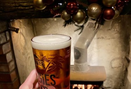 One hand holding up a glass full with beer, festive fireplace and decorations shown in background