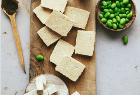 Chopping board on bench top with thick slices of tofu scattered and a small side pot of edamame beans