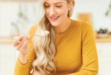 Dr Megan Rossi wearing a yellow shirt, sitting in front of a bowl of porridge while lifting a spoon to her mouth