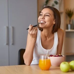 An image of a women eating and smiling. She has a bowl of food in front of her, orange juice and an apple.