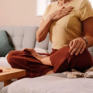 A photo of a woman sitting on a couch with her legs crossed, hand on her chest, doing a breathing exercise