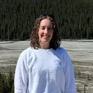 A photo of Lauren smiling. She has brunette, curly, mid-length hair and is stood in front of a vast landscape of trees.