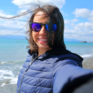 A photo of a woman with dark hair and sunglasses smiling. She's at the beach and the wind is blowing her hair.