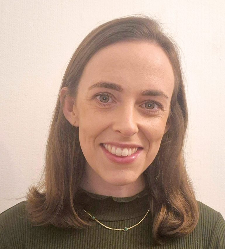 A photo of Alice Twomey smiling, she has brunette, shoulder length hair and green eyes