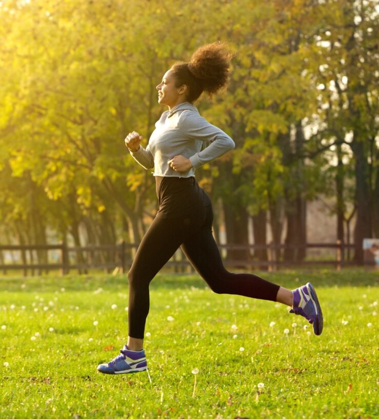 A photo of a woman running outside with trees in the background and sun shining down